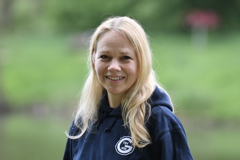 L&auml;chelnde Frau mit l&auml;ngeren blonden Haaren, in einem blauen Hoodie, steht vor einer gr&uuml;nen Naturlandschaft.