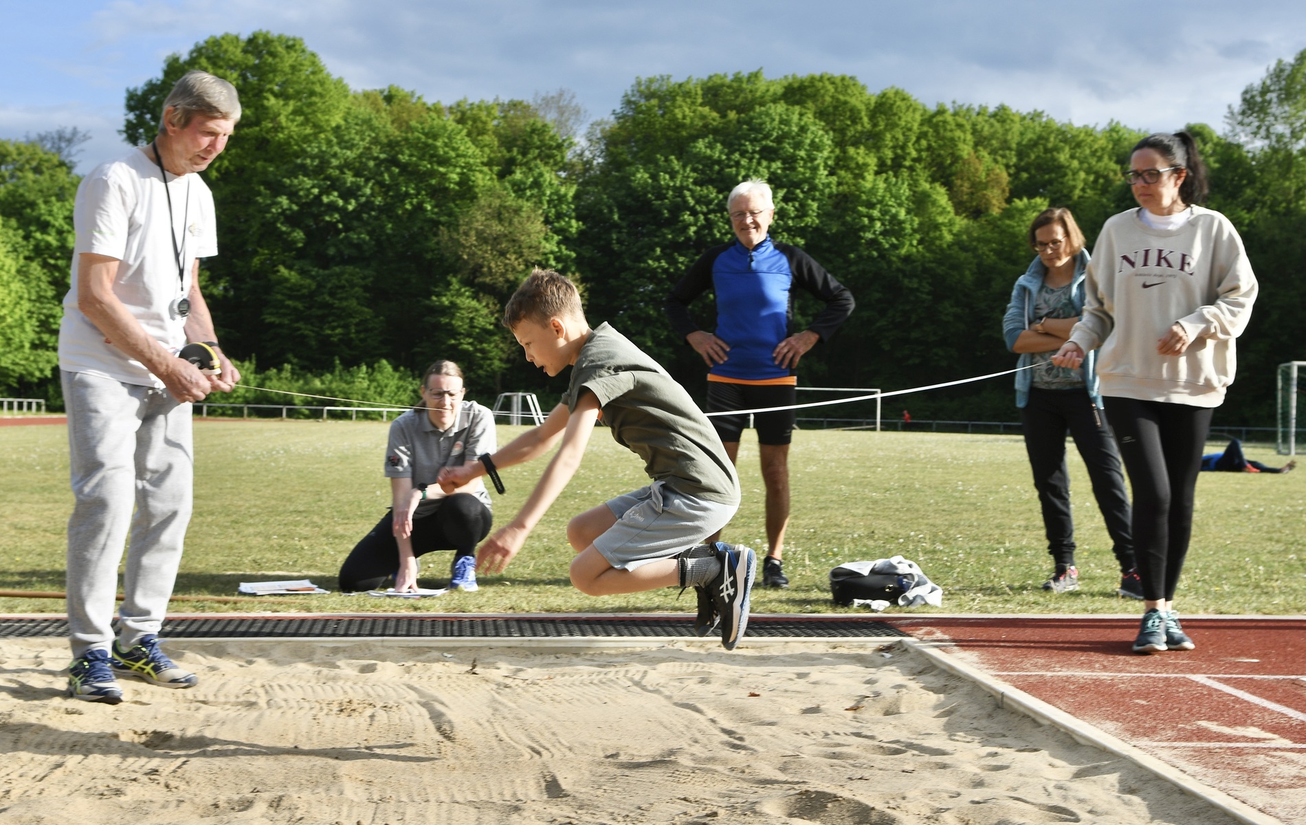 Junge springt im Weitsprungtraining auf einer Sandfl&auml;che, w&auml;hrend Trainer und Zuschauer im Hintergrund beobachten.