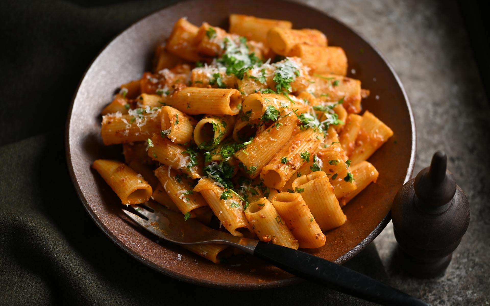 Teller mit rigatoni in Tomatensoße, garniert mit Kräutern und Parmesan, neben einer Gabel und Salzstreuer.