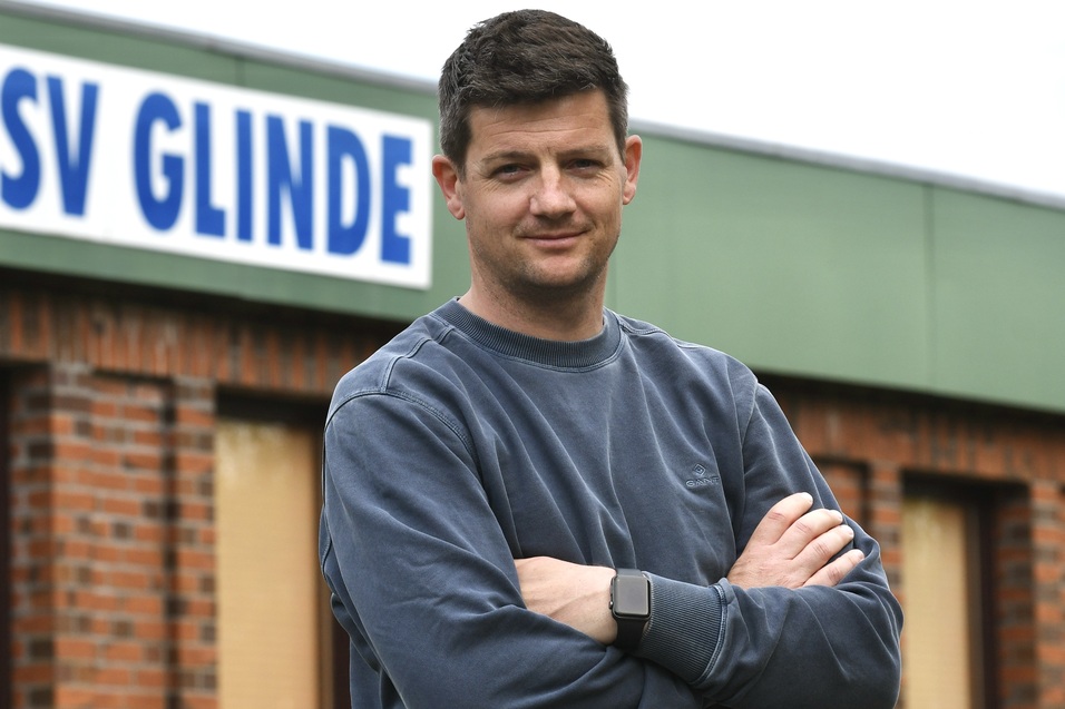 Mann mit verschr&auml;nkten Armen steht vor einem Geb&auml;ude mit dem Schriftzug &bdquo;TSV Glinde&ldquo; im Hintergrund.