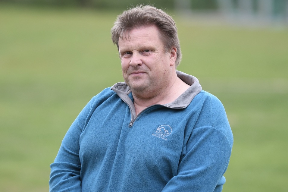 Ein Mann mit hellem Pullover steht im Freien vor einer gr&uuml;nen Wiese.