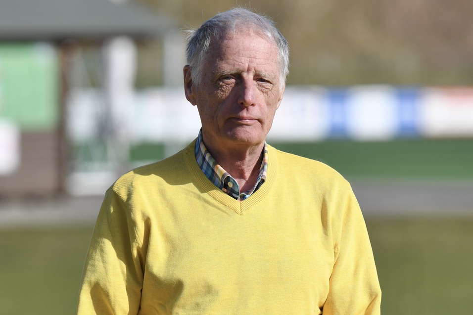 &Auml;lterer Mann in einem gelben Pullover steht im Freien vor einer gr&uuml;nen Sportanlage. Sonnenschein und klare Sicht.