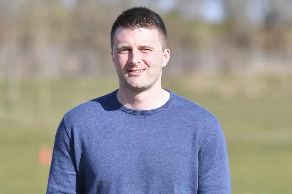 Mann mit kurzem Haar in blauer Kleidung steht l&auml;chelnd auf einem gr&uuml;nen Sportplatz im Freien.