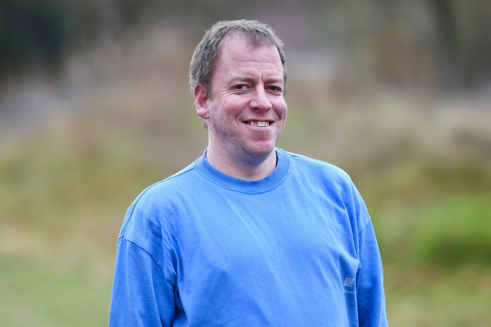 Ein Mann l&auml;chelt freundlich in einem blauen Sweatshirt vor unscharfem, nat&uuml;rlichem Hintergrund.