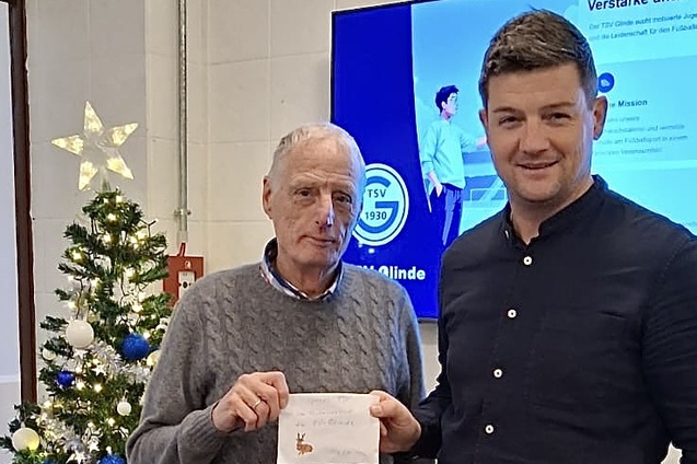 Zwei M&auml;nner stehen nebeneinander, einer h&auml;lt einen Umschlag. Im Hintergrund ein Weihnachtsbaum und ein Bildschirm.