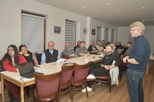Gruppe von Menschen sitzt an Tischen in einem Raum, einer spricht, w&auml;hrend andere aufmerksam zuh&ouml;ren.