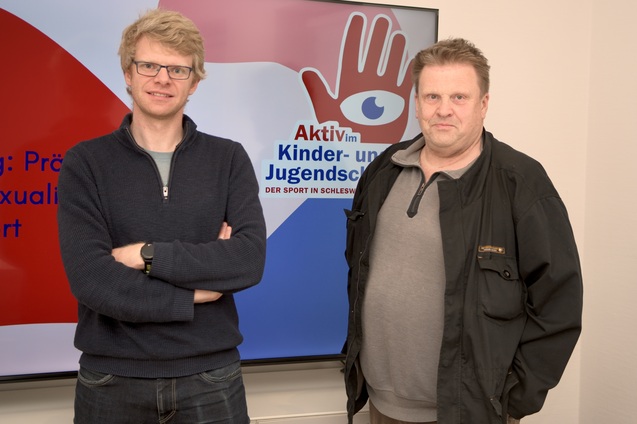 Zwei M&auml;nner stehen vor einem Bildschirm mit dem Schriftzug &bdquo;Aktiv im Kinder- und Jugendsport&ldquo; und einem Logo.