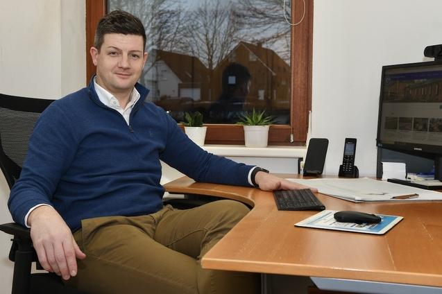 Mann sitzt an einem Schreibtisch, l&auml;chelt, vor ihm ein Computer und B&uuml;ropflanzen, Fenster im Hintergrund.