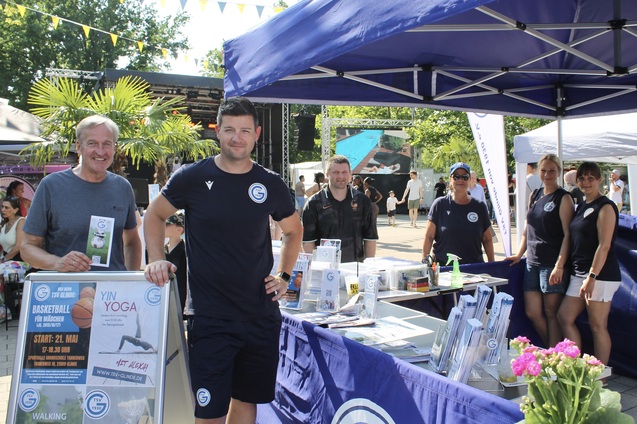 M&auml;nner und Frauen stehen am Info-Stand eines Sportevents mit Flyern und Werbematerialien im Freien.