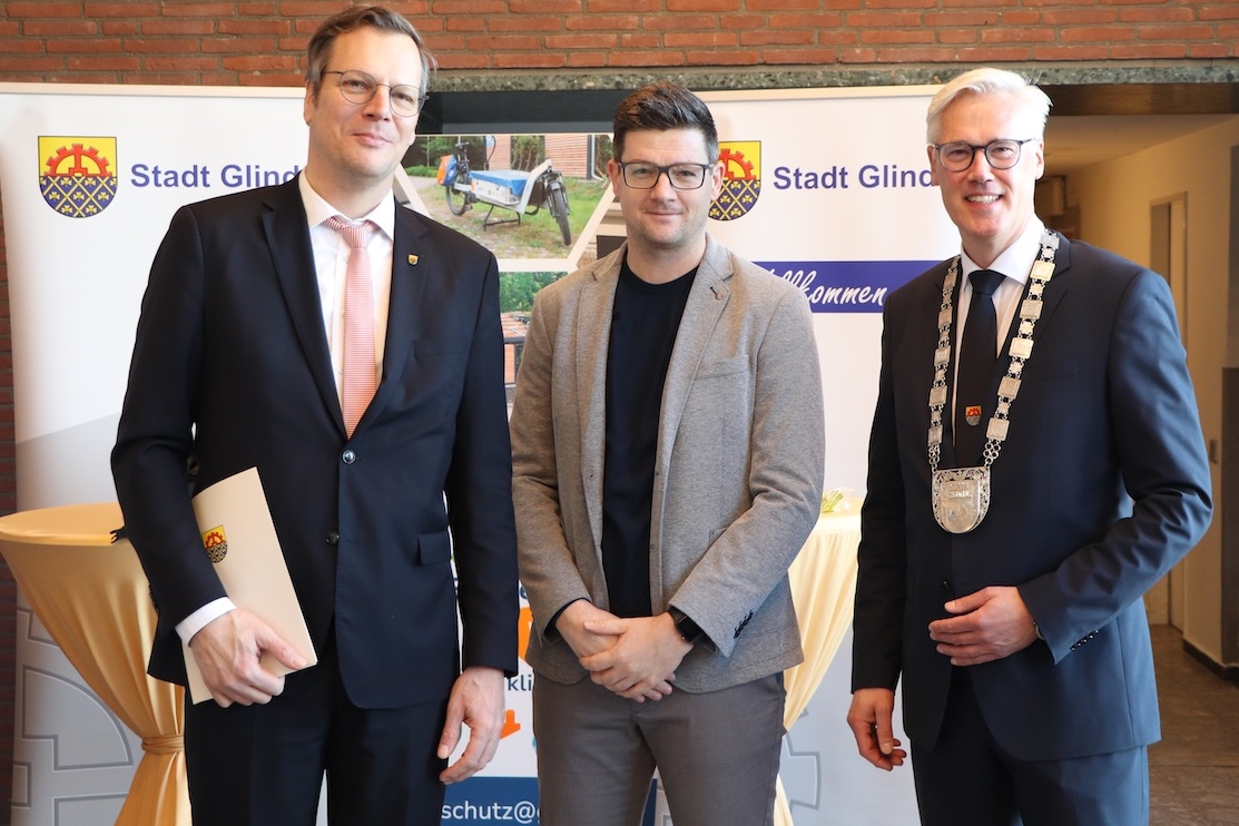Drei M&auml;nner in formeller Kleidung stehen vor einem Banner der Stadt Glinde bei einer Veranstaltung.