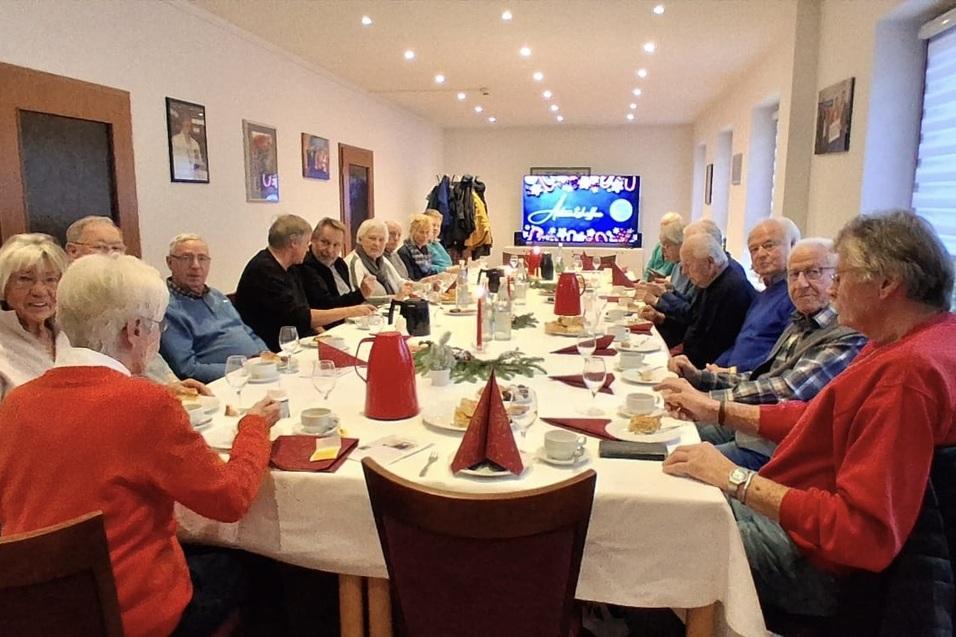 Gruppe älterer Menschen sitzt an festlich gedecktem Tisch in einem Raum mit Fernseher im Hintergrund.