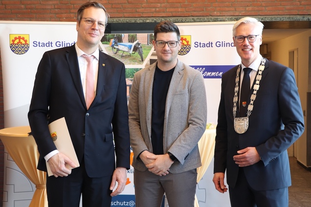 Drei M&auml;nner in formeller Kleidung stehen vor einem Banner der Stadt Glinde bei einer Veranstaltung.