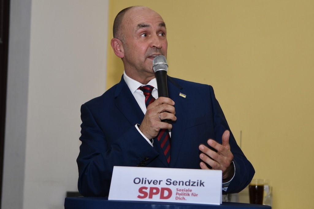 Mann im Anzug spricht mit Mikrofon, steht vor einem Tisch mit einem Schild &bdquo;Oliver Sendzik SPD&ldquo;. Hintergrund gelb.