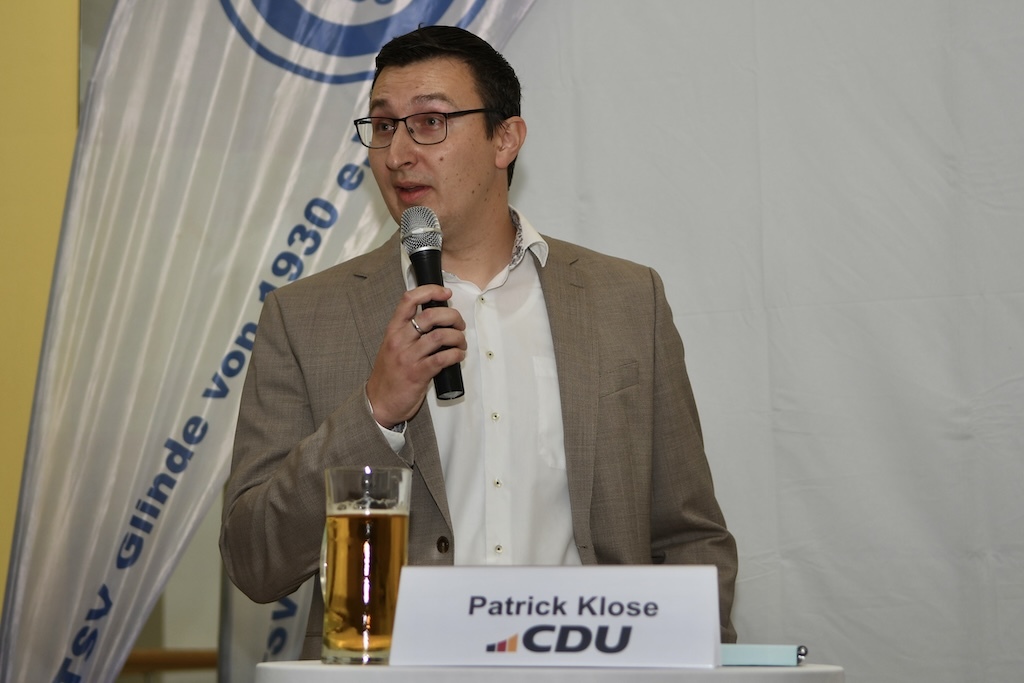 Ein Mann spricht mit Mikrofon, sitzt am Tisch mit Bierglas und Schild mit seinem Namen und CDU-Logo im Hintergrund.