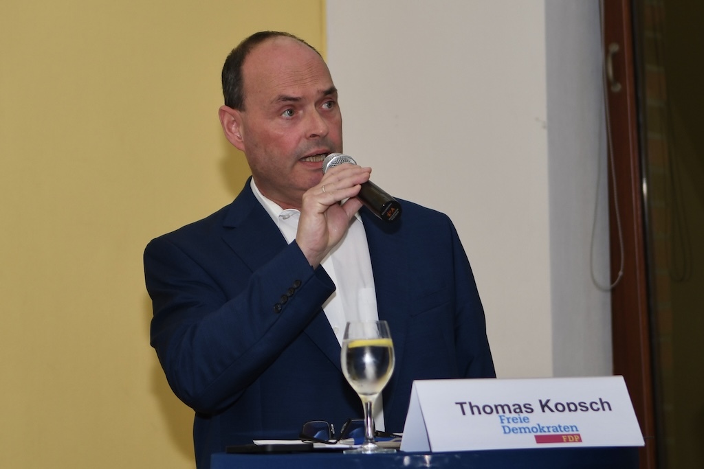 Mann mit Mikrofon spricht vor Publikum, steht an einem Tisch mit Glas Wasser und Namensschild &bdquo;Thomas Koosch, Freie Demokraten&ldquo;.