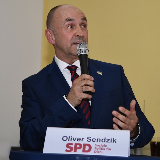 Mann im Anzug spricht mit Mikrofon, steht vor einem Tisch mit einem Schild &bdquo;Oliver Sendzik SPD&ldquo;. Hintergrund gelb.