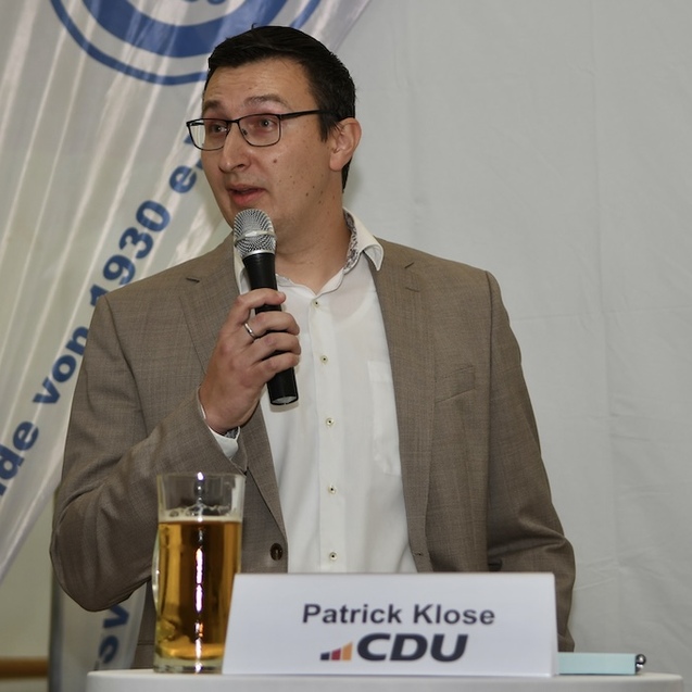 Ein Mann spricht mit Mikrofon, sitzt am Tisch mit Bierglas und Schild mit seinem Namen und CDU-Logo im Hintergrund.