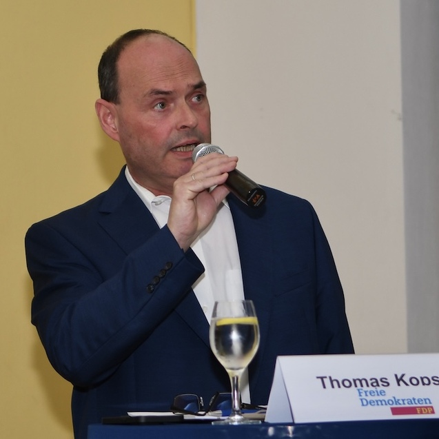Mann mit Mikrofon spricht vor Publikum, steht an einem Tisch mit Glas Wasser und Namensschild &bdquo;Thomas Koosch, Freie Demokraten&ldquo;.