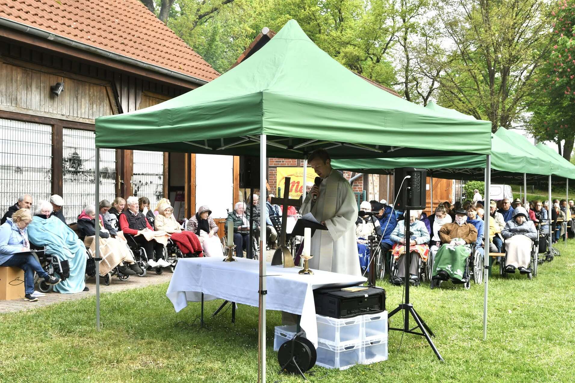 Pfarrer hält eine Ansprache unter einem grünen Zelt vor einer versammelten Gruppe älterer Menschen in Rollstühlen.