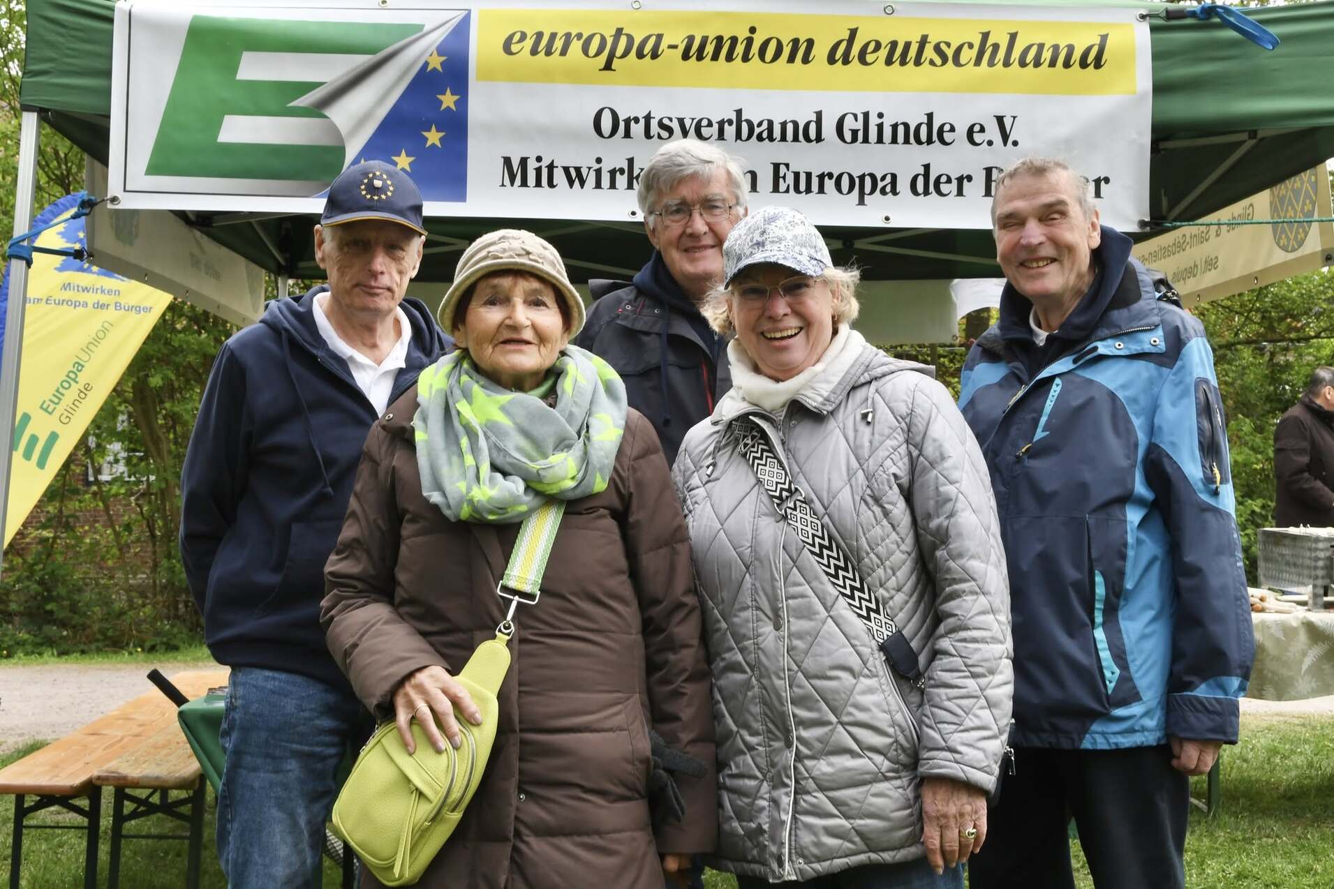 Fünf Personen stehen lächelnd vor einem Zeltdach mit dem Banner „europa-union deutschland“ in einem Park.