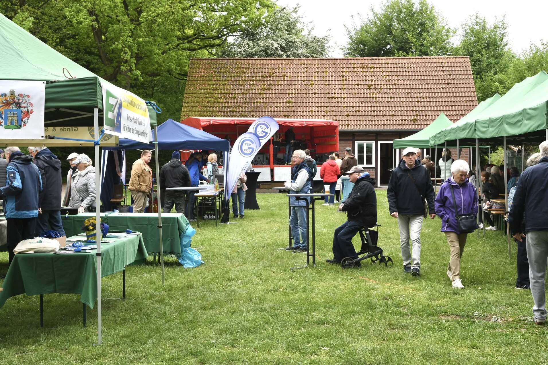Menschen besuchen einen Outdoor-Markt mit Ständen unter grünen Zelten in einer Gartenlandschaft.