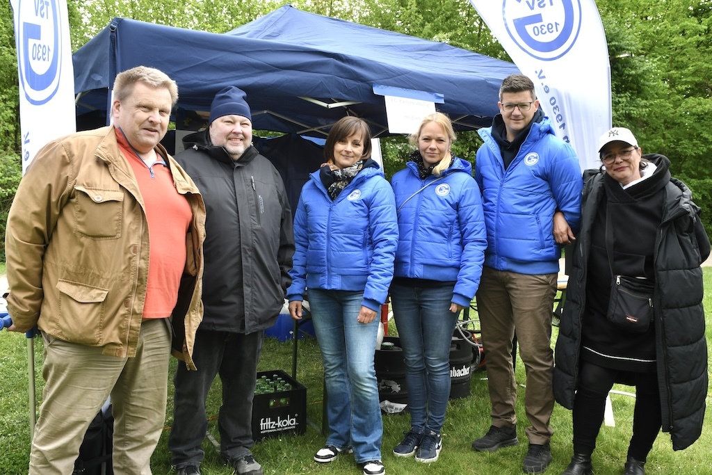 Sechs Personen in blauen Jacken stehen vor einem Pavillon im Freien, umgeben von gr&uuml;ner Natur.