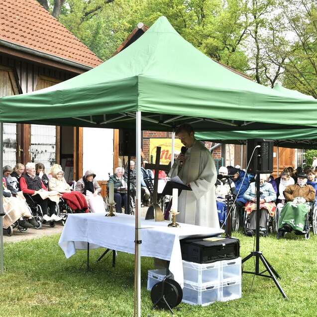 Pfarrer hält eine Ansprache unter einem grünen Zelt vor einer versammelten Gruppe älterer Menschen in Rollstühlen.