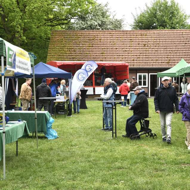 Menschen besuchen einen Outdoor-Markt mit Ständen unter grünen Zelten in einer Gartenlandschaft.