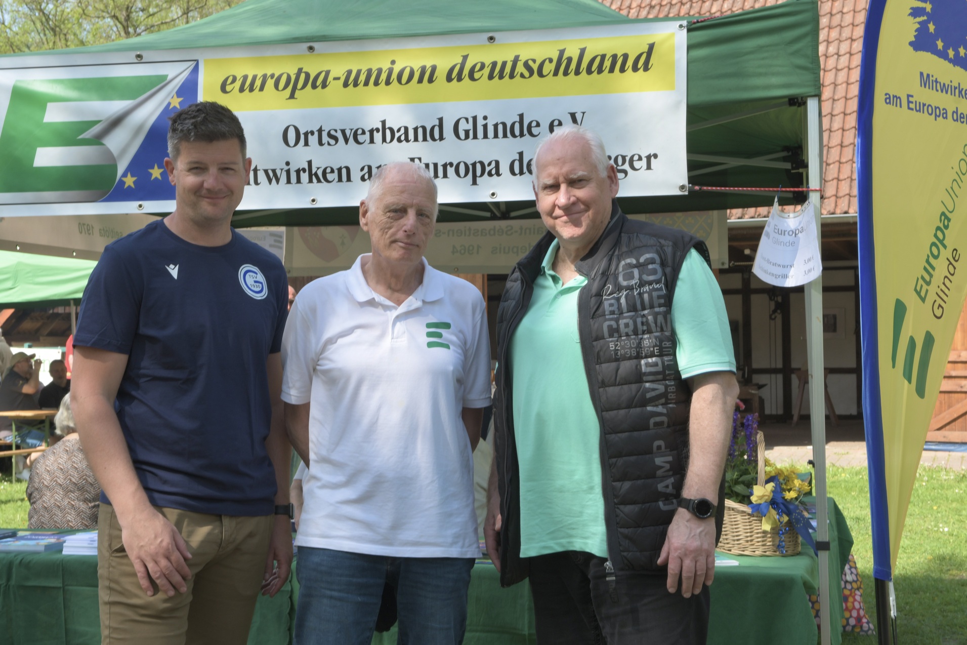 Drei M&auml;nner stehen l&auml;chelnd vor einem Stand der Europa-Union Deutschland, bemerkt durch ein gr&uuml;nes Banner und Tische.
