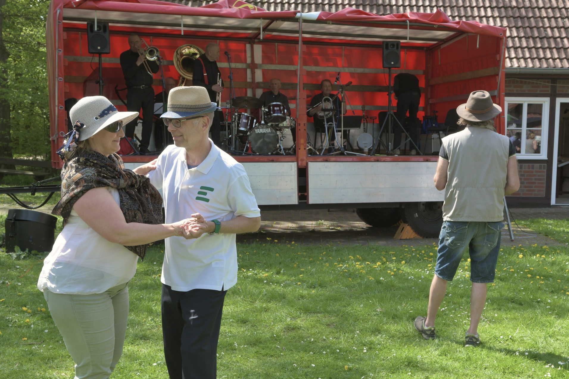 Tanzendes Paar vor einer B&uuml;hne mit einer Jazzband, umgeben von Wiese und B&auml;umen an einem sonnigen Tag.