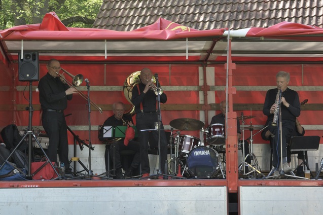 Eine Band mit sieben Musikern spielt auf einer Roten B&uuml;hne, umgeben von Instrumenten wie Trompeten, Posaune und Schlagzeug.