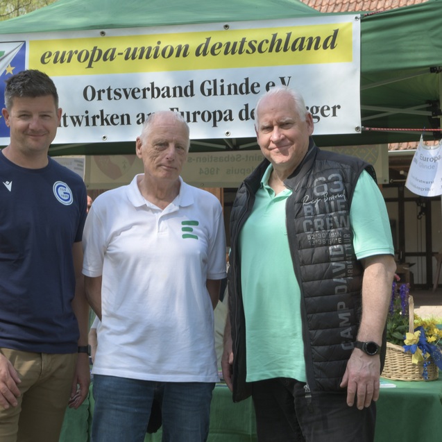 Drei M&auml;nner stehen l&auml;chelnd vor einem Stand der Europa-Union Deutschland, bemerkt durch ein gr&uuml;nes Banner und Tische.