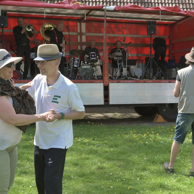 Tanzendes Paar vor einer B&uuml;hne mit einer Jazzband, umgeben von Wiese und B&auml;umen an einem sonnigen Tag.