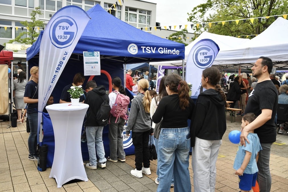 Menschen stehen Schlange vor einem Informationsstand des TSV Glinde auf einem belebten Platz.