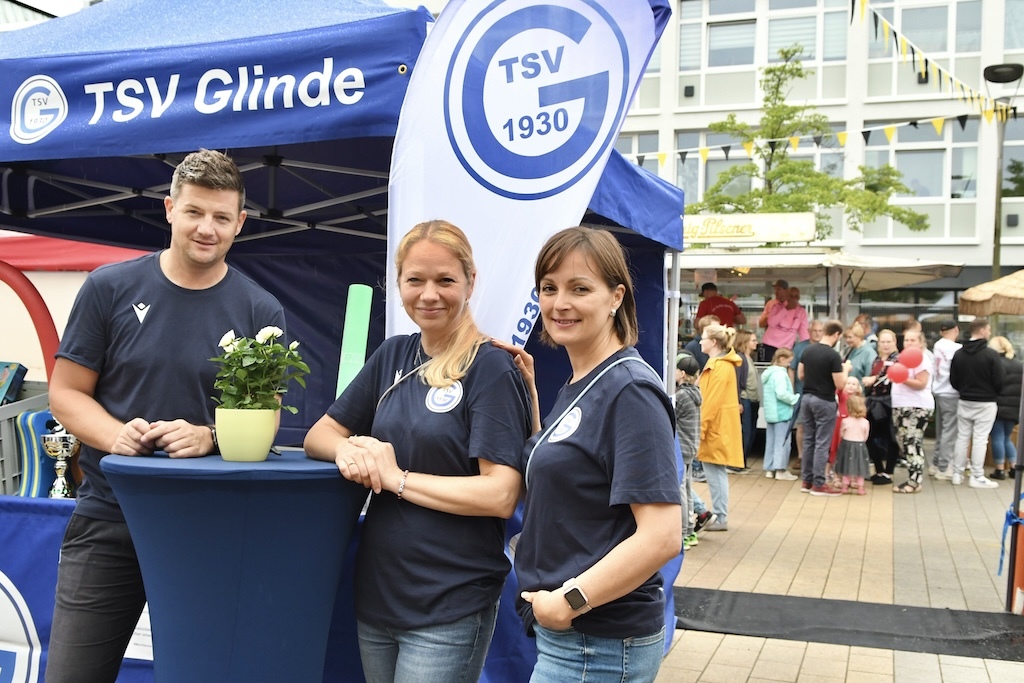 Drei Personen inTSV Glinde-T-Shirts stehen an einem Stehtisch vor einem blauen Vereinsstand auf einer Veranstaltung.