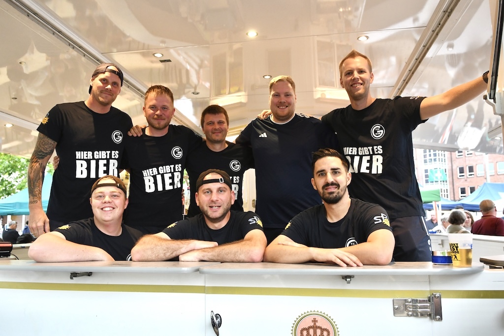 Gruppe von acht Personen in schwarzen T-Shirts hinter einem Bierstand mit dem Schriftzug &bdquo;Hier gibt es Bier&ldquo;.