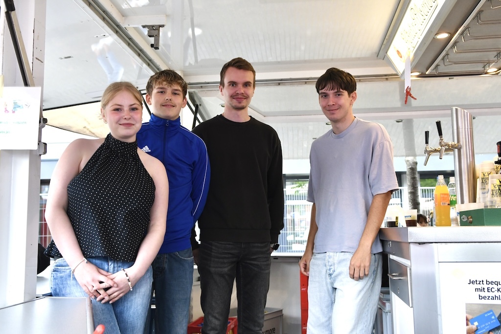 Vier junge Leute stehen in einem Verkaufsstand, mit Getr&auml;nken im Hintergrund und l&auml;chelnd in die Kamera blickend.