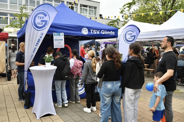 Menschen stehen Schlange vor einem Informationsstand des TSV Glinde auf einem belebten Platz.