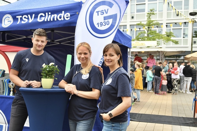 Drei Personen inTSV Glinde-T-Shirts stehen an einem Stehtisch vor einem blauen Vereinsstand auf einer Veranstaltung.