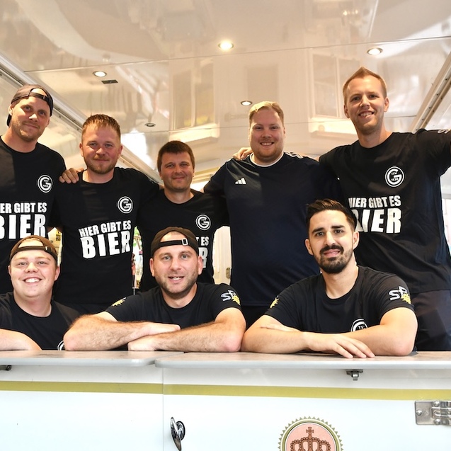 Gruppe von acht Personen in schwarzen T-Shirts hinter einem Bierstand mit dem Schriftzug &bdquo;Hier gibt es Bier&ldquo;.