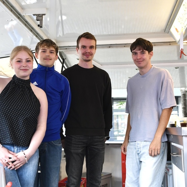 Vier junge Leute stehen in einem Verkaufsstand, mit Getr&auml;nken im Hintergrund und l&auml;chelnd in die Kamera blickend.