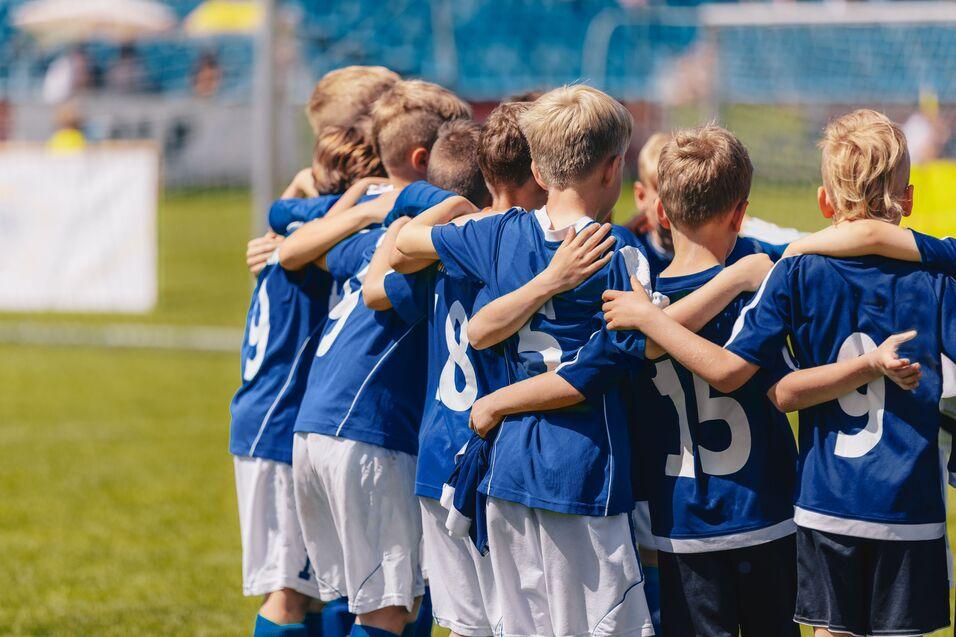 Gruppe von Kindern in blauen Fu&szlig;balltrikots, die sich lachend umarmen und auf einem Sportplatz stehen.