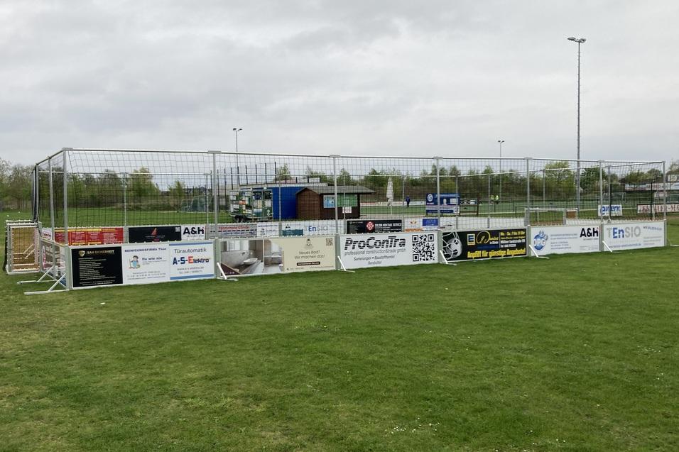 Umzäunte Fläche auf einem Rasenplatz mit sichtbaren Werbebannern, im Hintergrund sind Sporteinrichtungen zu erkennen.