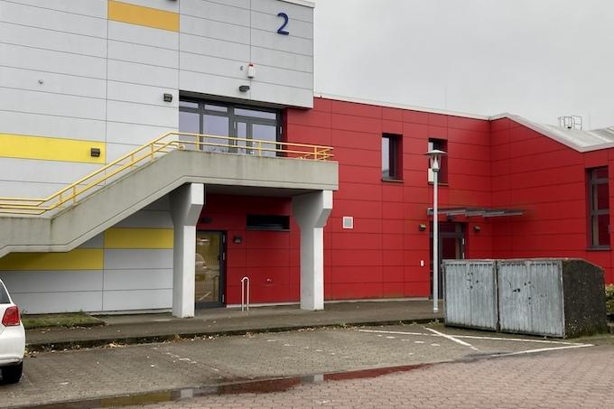 Fassade eines modernen Gebäudes in rot und grau mit Stufen, einem Pkw und Abfallcontainer im Vordergrund.