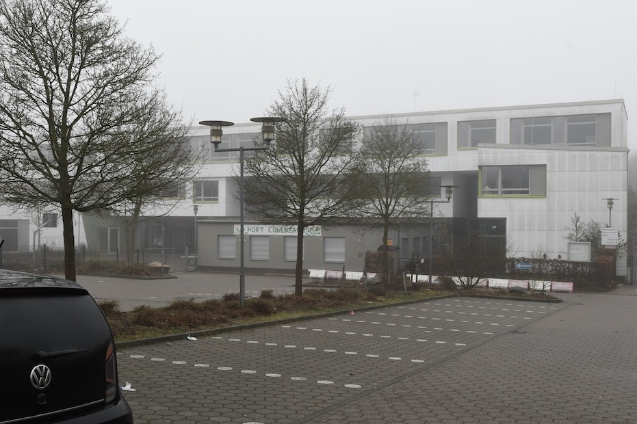 Moderne Schule im Nebel mit einem Parkplatz, B&auml;umen im Vordergrund und einem schwarzen VW PKW.