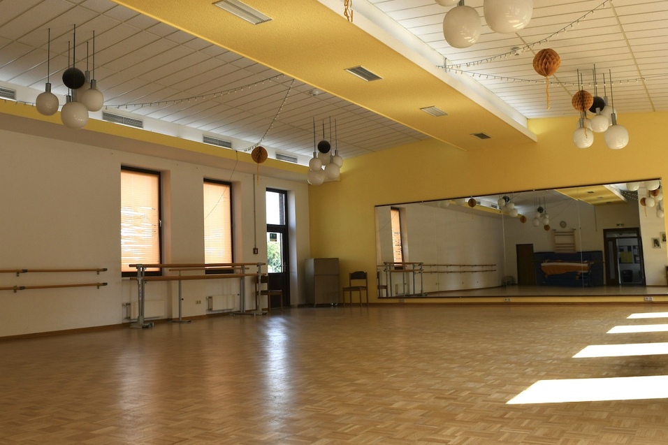 Tanzstudio mit Parkettboden, gro&szlig;en Fenstern und Spiegelwand, beleuchtet von runden Deckenlampen.