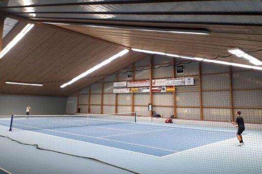 Zwei Tennisspieler auf einem blauen Platz in einer Halle, umgeben von einem Netz und Holzwänden.