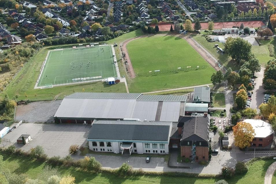 Luftaufnahme eines Sportgeländes mit Fußballfeld, mehreren Gebäuden und Wohngebieten im Hintergrund.