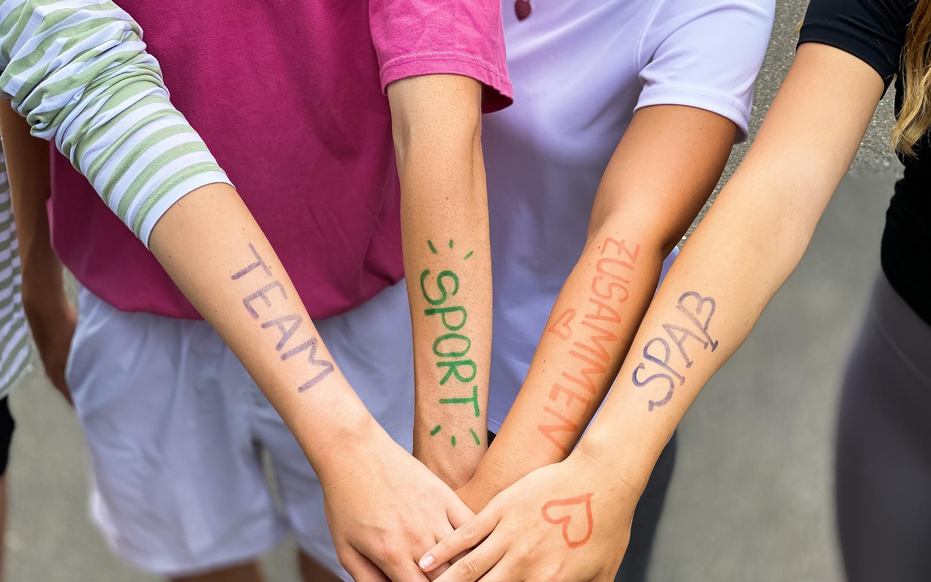 Vier Arme liegen übereinander, mit farbiger Schrift: „TEAM“, „SPORT“, „ZUSAMMEN“, „SPASS“ auf der Haut.