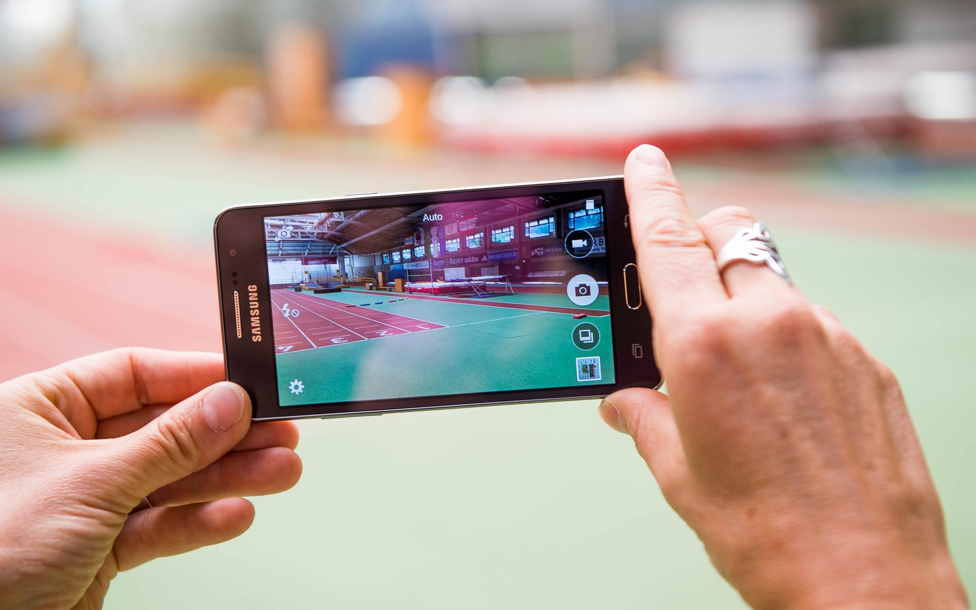 Hand hält ein Smartphone, das eine Sporthalle mit einer Laufbahn im Hintergrund zeigt.